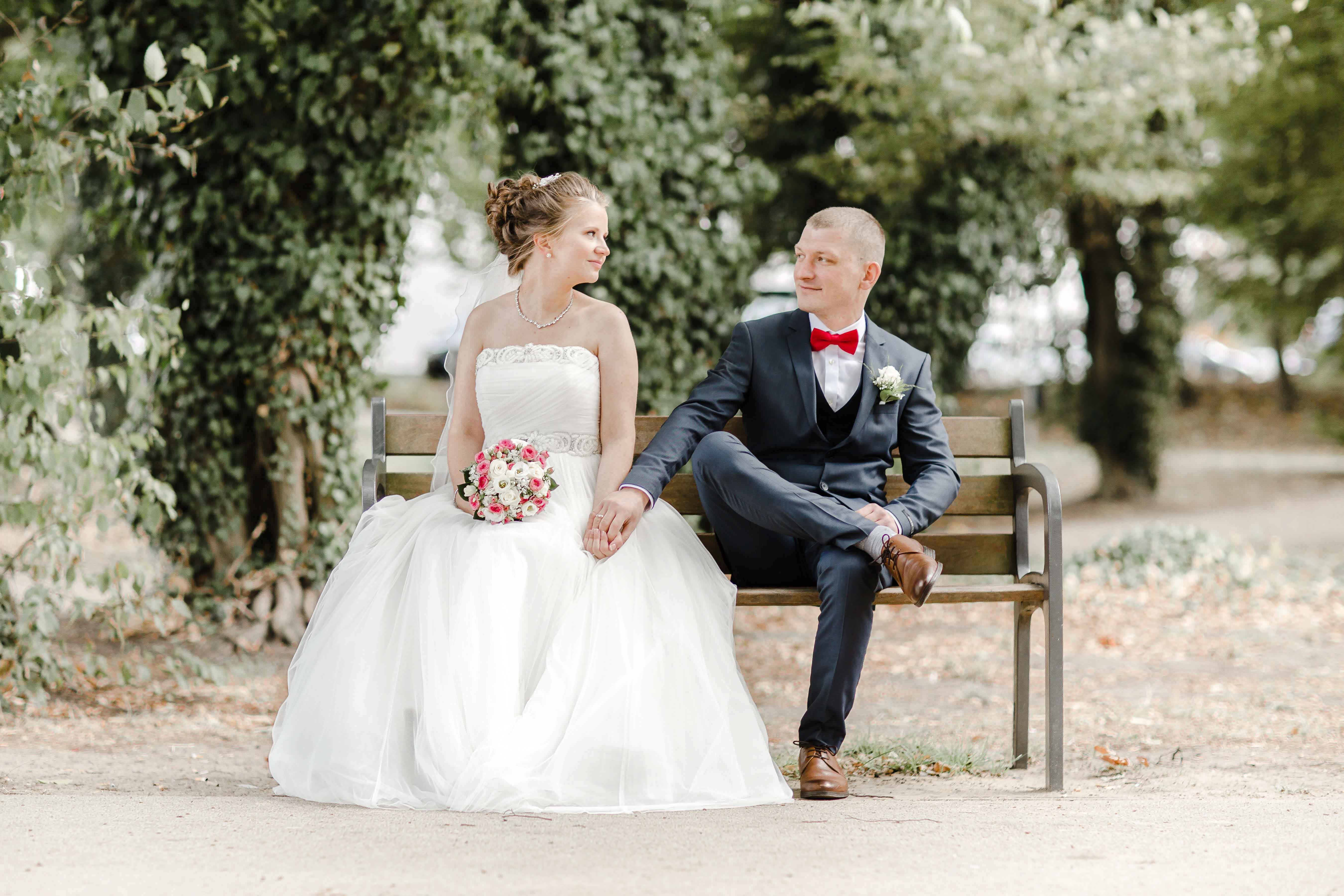 Hochzeitspaar sitzt auf Parkbank und lächelt sich an